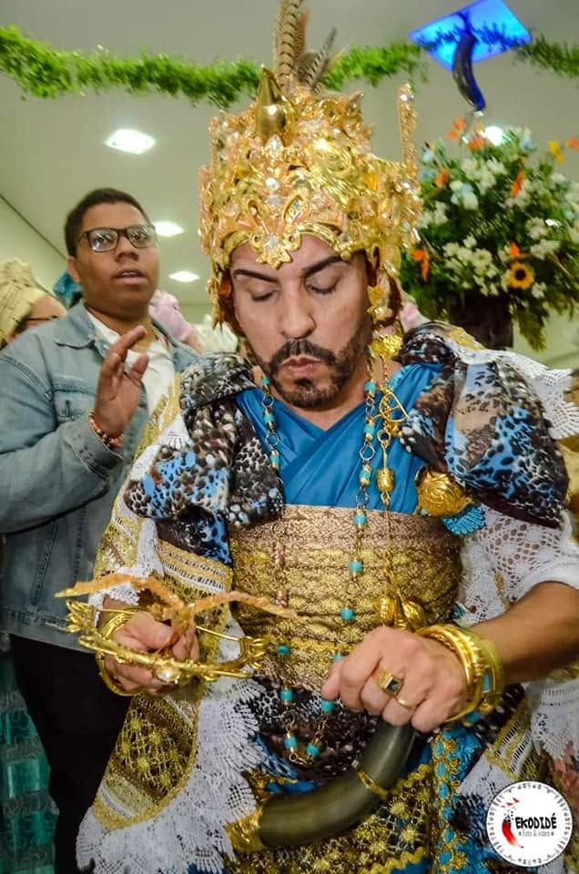 Pai Diney paramentado de Ọ̀ṣọ́ọ̀sì, com coroa dourada, ojá azul e adê de metal, em dia de festa